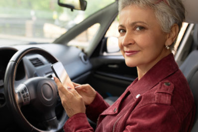 Mulher branca idosa, com camisa vinho, segurando um celular sentada ao volante do carro