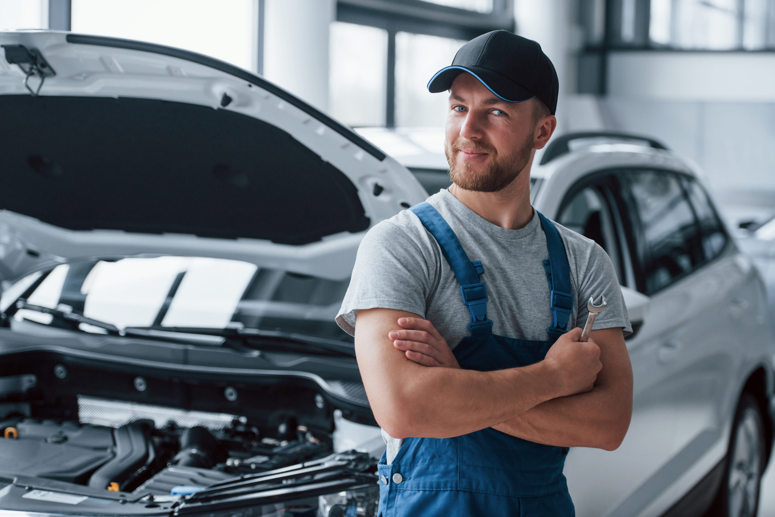 Mecânico branco usando macacão azul sorri encostado em um carro com o capô aberto