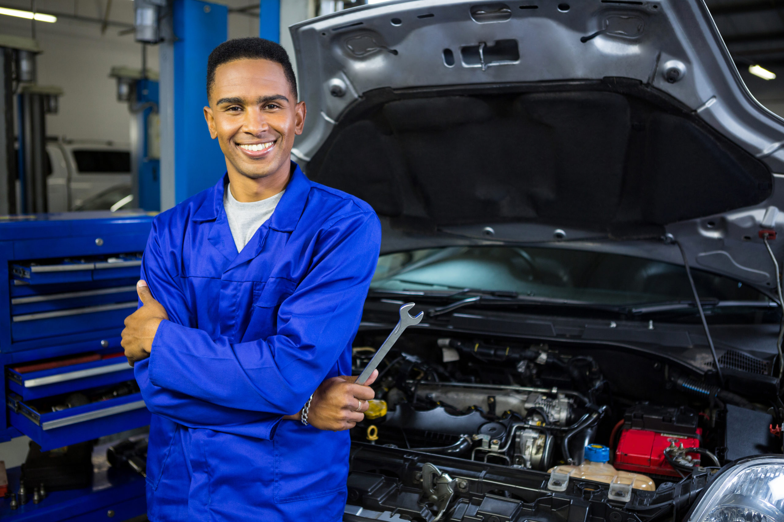 Mecânico negro usando um macacão azul e segurando uma chave de fenda sorrindo para a câmera encostado em um carro com o capô aberto