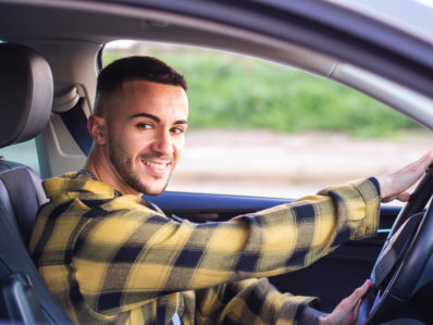 Homem branco, com camisa amarela com preto, sorrindo para a câmera ao volante de um carro