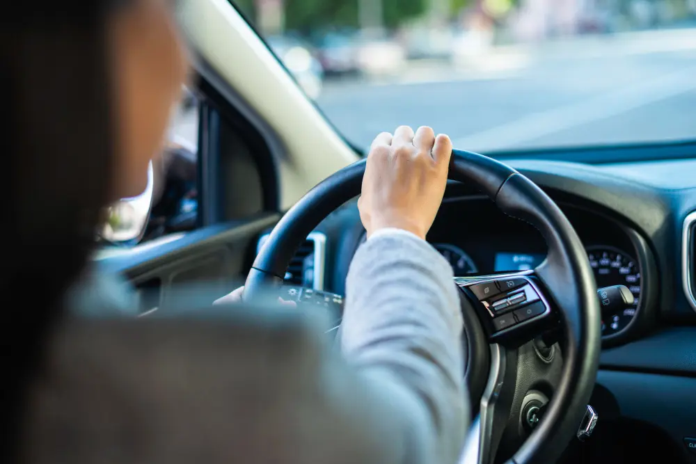 Visão de costas de uma pessoa branca, com camisa de mangas compridas, com a mão direita no volante do carro