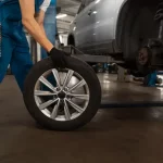Mecânico branco com macacão azul manuseando um pneu para colocá-lo no carro