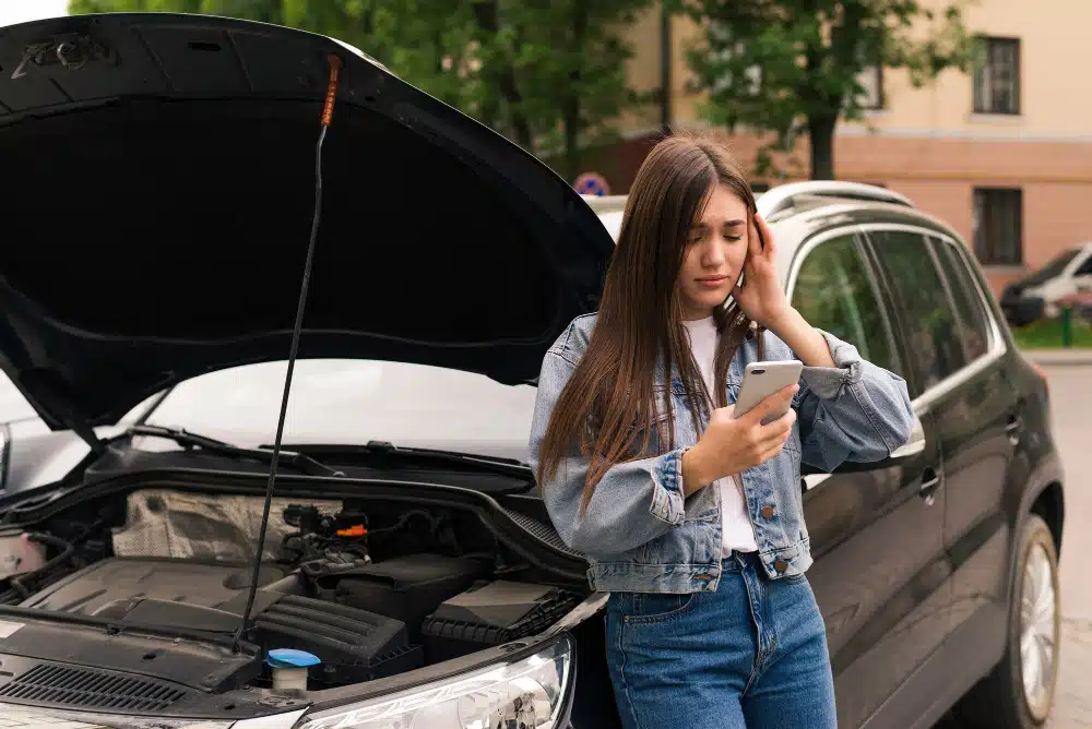 Mulher branca, de cabelo liso e longo, com jaqueta jeans, olha triste para o celular encostada no carro com o capô aberto
