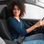 Mulher negra com blusa azul e jeans sentada ao volante do carro olhando para a pessoa no banco do carona