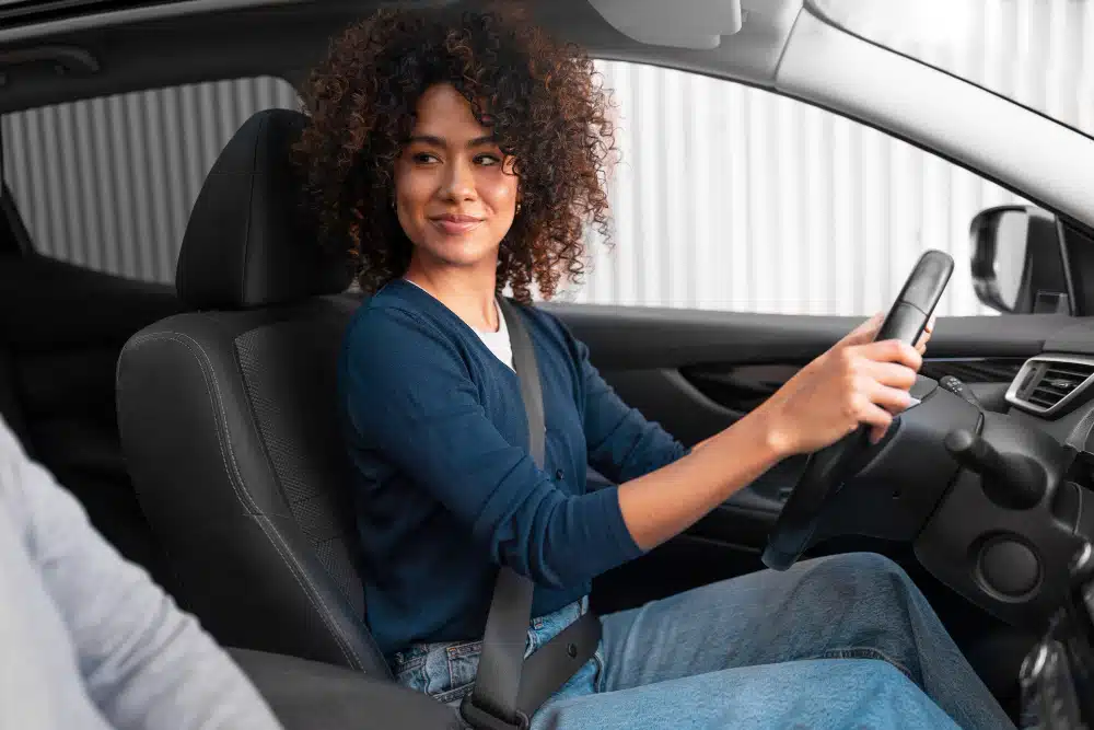 Mulher negra com blusa azul e jeans sentada ao volante do carro olhando para a pessoa no banco do carona