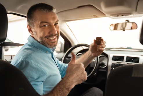 Homem branco ao volante de um carro, com camisa azul, fazendo sinal de positivo com a mão e sorrindo para a câmera que está atrás dele