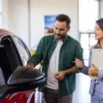 Homem branco em loja de carros com a vendedora ao lado olhando um carro vermelho