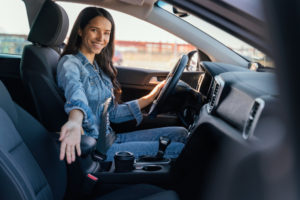 Mulher branca ao volante de um carro, com roupa jeans, sorri para a câmera com o braço direito estendido