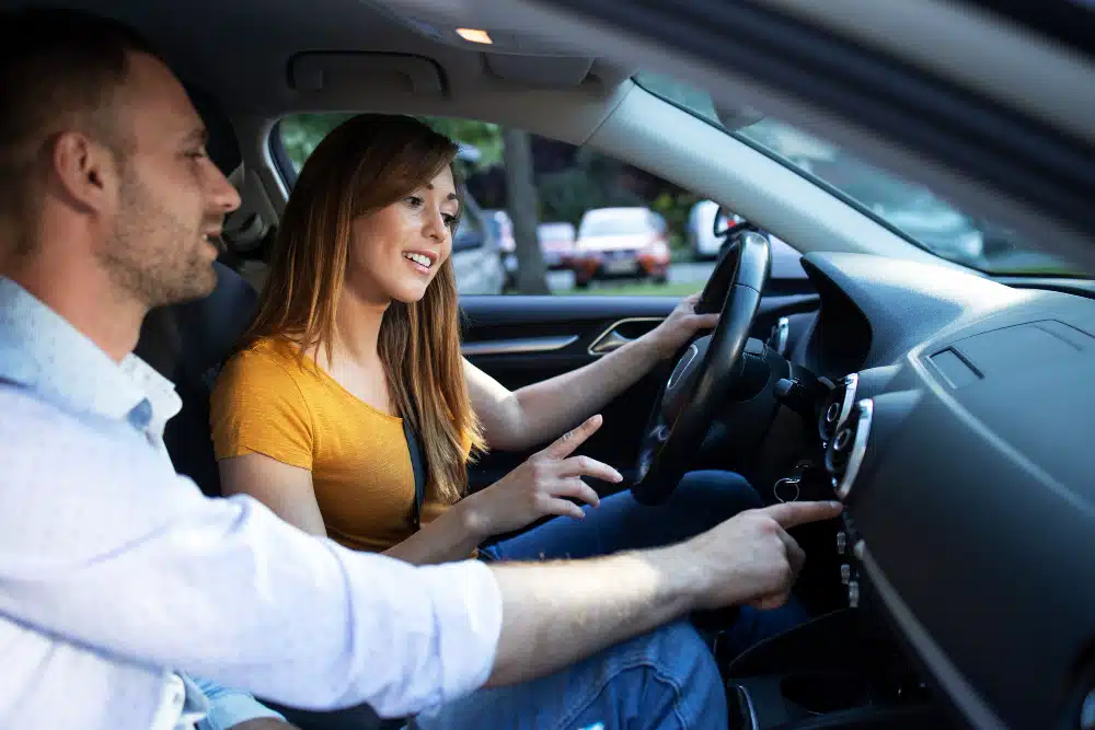 Mulher branca de cabelo iso e camiseta amarela ao volante de um carro com um homem branco no banco do carona apontando para o painel